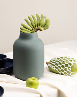 Eine grüne Vase mit Bananen steht auf einem Tisch neben einem Apfel und einer verpackten Papaya.