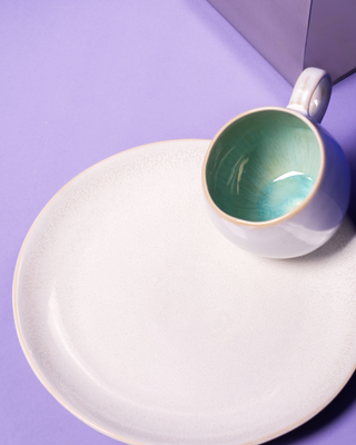 Weiße Tasse mit hellgrünem Inneren und passendem Teller auf einem lilafarbenen Hintergrund.