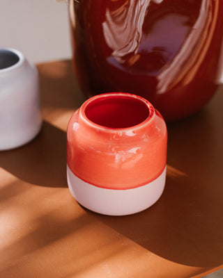 Kleine bunte Vase mit rotem und weißem Design steht auf einem braunen Tisch, umgeben von weiteren Tonkrügen in sanftem Licht.
