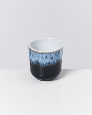 Blau und schwarz glasierte Keramiktasse vor einem hellgrauen Hintergrund.