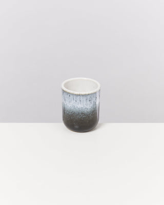Keramiktasse mit einem Farbverlauf von Schwarz zu Weiß auf einem weißen Tisch vor grauem Hintergrund.