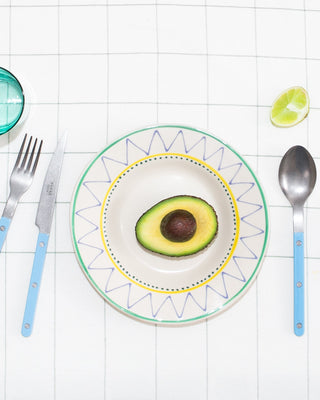 Ein Teller mit halber Avocado, Messer, Gabel und Löffel auf kariertem Tisch, daneben eine Limettenscheibe.