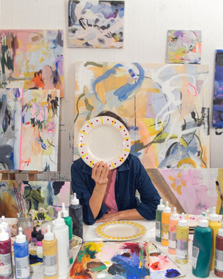 Eine Person hält einen bemalten Teller vor das Gesicht, umgeben von abstrakten Gemälden und Farbflaschen im Atelier.