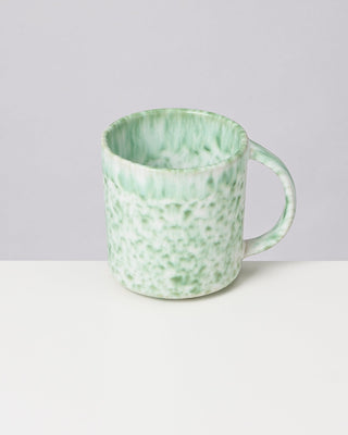 Grüne, gesprenkelte Keramiktasse mit Henkel auf weißem Tisch vor grauem Hintergrund.