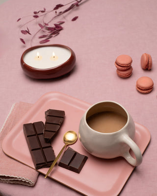 Ein Tisch mit Kerze, Schokolade, einer Tasse Kaffee, einem goldenen Löffel und rosafarbenen Macarons auf rosa Tischdecke.