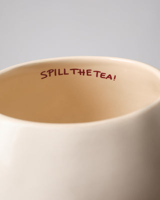 Beige Tasse mit der Aufschrift "SPILL THE TEA!" innen, vor einem grauen Hintergrund.