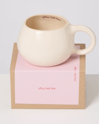 Beiger Keramikbecher mit der Aufschrift „Spill the Tea!“ auf einem pinken Karton mit gleichem Text.