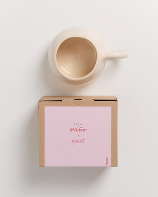 Beiger Keramikbecher und brauner Karton mit rosa Etikett, auf dem "Motel a Mio x Ankat" steht.