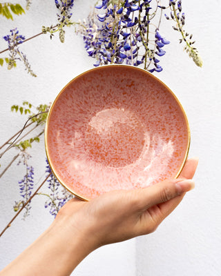 Eine Hand hält eine rosafarbene Schale vor lila Blumen vor einer weißen Wand.