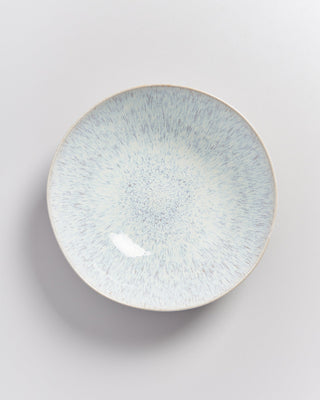 Ein runder, weißer Teller mit blauen Sprenkeln und einem leicht glasierten Finish auf einer hellgrauen Oberfläche.