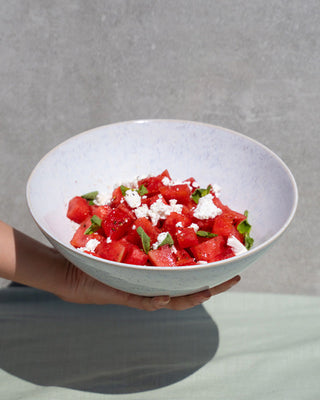 Weiße Schüssel mit Wassermelonensalat, Feta, Minze und einer Hand, die sie hält, vor grauem Hintergrund.