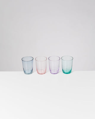 Vier bunte Gläser in Blau, Rosa, Lila und Grün stehen auf einer weißen Oberfläche vor hellem Hintergrund.
