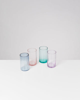Vier bunte trinkgläser in Blau, Rosa, Grün und Lila auf einem weißen Tisch vor einem grauen Hintergrund.