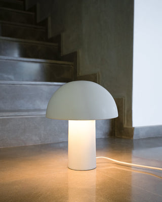 Weiße Tischlampe mit pilzförmigem Schirm leuchtet auf reflektierendem Boden neben einer Treppe.