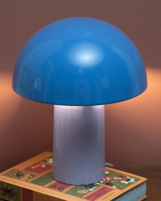 Blaue Pilzlampe auf einem Buch mit farbigem Cover, vor einer braunen Wand.