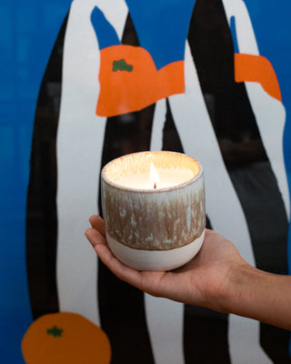 Eine Hand hält eine brennende Kerze vor einem abstrakten, bunten Hintergrund mit orangenen und schwarzen Formen.