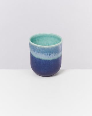Blauer Keramikbecher mit grünem Innenleben steht auf einem weißen Tisch vor einer grauen Wand.