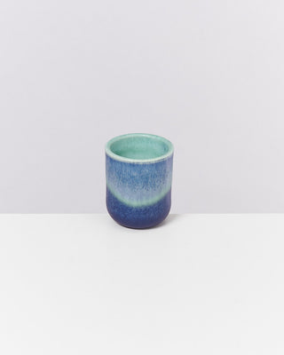 Kleiner Becher in blauem und türkisfarbenem Farbverlauf auf einem weißen Tisch vor hellem Hintergrund.