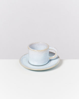 Graue Keramiktasse mit Untertasse auf einem weißen Tisch vor einem grauen Hintergrund.