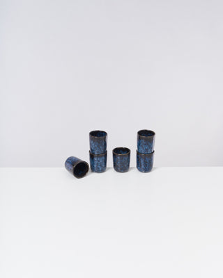 Sechs kleine, schwarze Becher mit blauen Mustern auf einem weißen Tisch; fünf stehend, einer liegend.