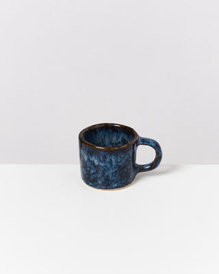 Eine blaue, keramikartige Tasse mit Henkel steht auf einer weißen Oberfläche vor einem neutralen Hintergrund.