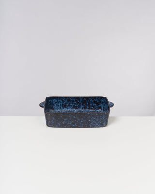 Blaue Keramikauflaufform mit Henkeln steht auf einem weißen Tisch vor neutralem Hintergrund.