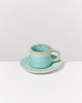Türkise Tasse mit Untertasse auf weißem Tisch vor grauem Hintergrund.