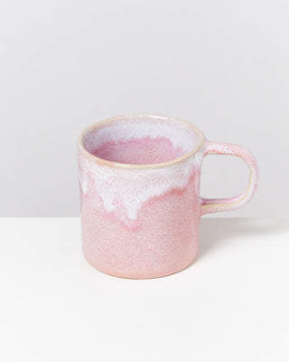 Eine rosa-weiße Keramiktasse mit Henkel steht auf einem weißen Untergrund vor einer grauen Wand.