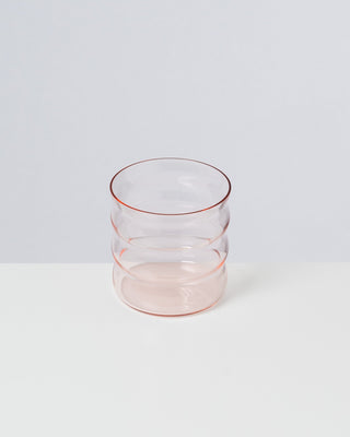 Ein durchsichtiges Glas mit gewellter Struktur steht auf einer weißen Oberfläche vor einem hellgrauen Hintergrund.