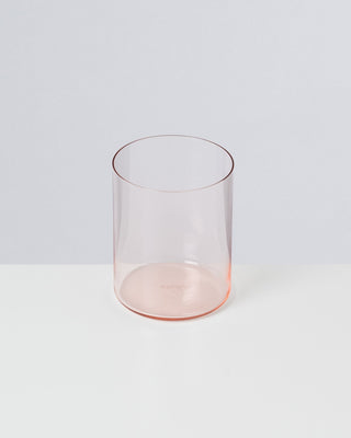 Ein rosa, transparentes Glas steht auf einer weißen Oberfläche vor einem grauen Hintergrund.