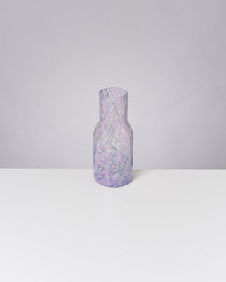 Ein gläserner, pastellfarbener Krug mit spiralförmigem Muster steht auf einem weißen Tisch vor hellgrauem Hintergrund.