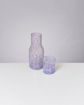 Lila und weiß gemusterte Glasflasche und -becher stehen auf einem weißen Tisch vor einem grauen Hintergrund.