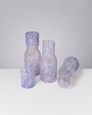 Mehrere Glasgefäße mit lila-blauem Muster stehen und liegen auf einem weißen Tisch vor grauem Hintergrund.