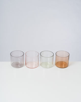 Vier farbige, durchsichtige Gläser in einer Reihe auf einem weißen Tisch vor grauem Hintergrund.