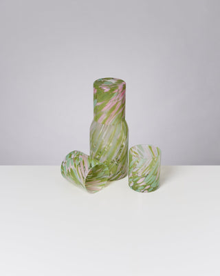 Glasflasche mit zwei farbigen Gläsern in Grün- und Rosatönen auf einem weißen Tisch.