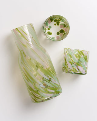 Glasflasche und zwei Trinkgläser mit grünem und rosa Muster auf weißem Hintergrund, von oben fotografiert.