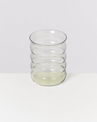 Ein klarer, wellenförmiger Glasbecher steht auf einem weißen Tisch vor einem neutralen Hintergrund.