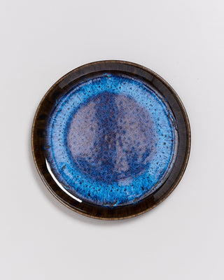 Blauer Keramikteller mit kunstvoller, gesprenkelter Glasur und braunem Rand auf weißem Hintergrund.