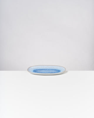 Ovale Keramikplatte mit blauer Mitte auf weißem Tisch vor grauem Hintergrund.