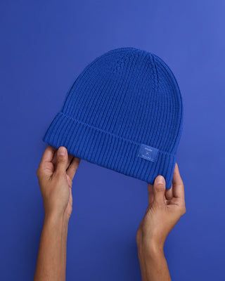 Hände halten eine blaue Strickmütze vor einem blauen Hintergrund.