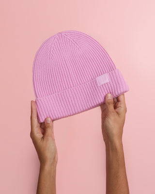 Hände halten eine rosa Strickmütze vor einem rosa Hintergrund.