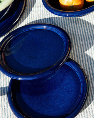 Zwei blaue, glänzende Keramikteller auf gestreiftem Stoff, neben einer Schale mit Orangen.