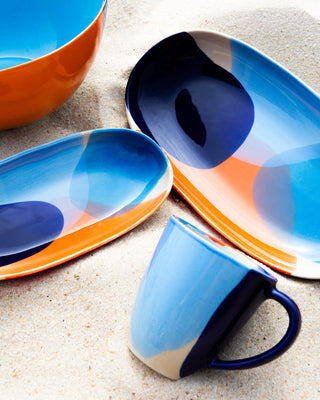Bunt bemaltes Geschirr und eine Tasse auf Sand liegend, mit blauen und orangefarbenen Mustern.