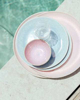Gestapelte Teller in Pastellrosa und Blau mit einem Glas, auf einer Steinoberfläche neben einem Pool.