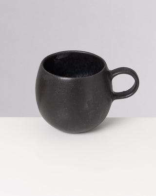 Schwarze, bauchige Keramiktasse mit Henkel auf einem weißen Tisch vor grauem Hintergrund.