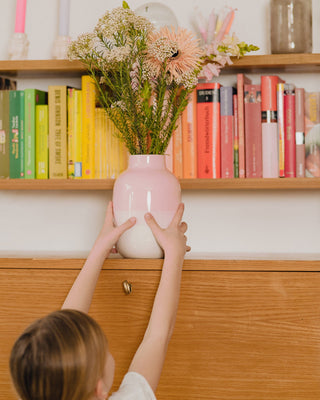 Ein Kind greift nach einer rosa Vase mit Blumen vor einem Regal voller bunter Bücher.