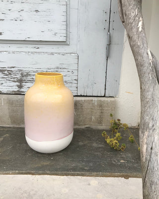 Eine große Vase in Gelb- und Rosatönen steht vor einer alten, blauen Tür neben einem vertrockneten Ast.