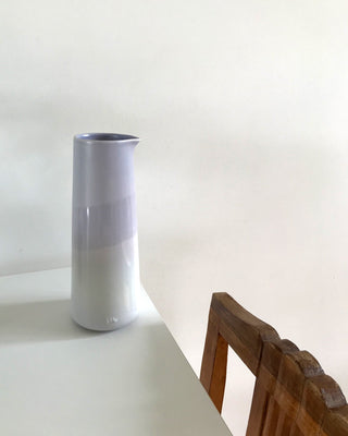 Eine hohe, zweifarbige Vase steht auf einem weißen Tisch neben einem hölzernen Stuhl vor einer weißen Wand.