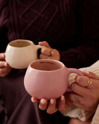 Zwei Personen halten Tassen, eine rosa und eine cremefarben, mit der Aufschrift "SPILL THE TEA!" am Rand.
