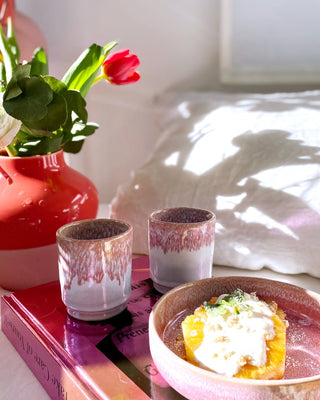 Pinkfarbener Krug mit Blume, zwei Kerzenhalter und Schale mit orangem Dessert auf einem Buchdeckel im Sonnenlicht.